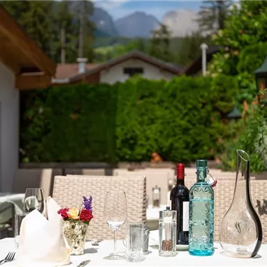 An outdoor table, beautifully set with glasses, wine bottles, and flowers. In the background, green bushes and mountains can be seen.