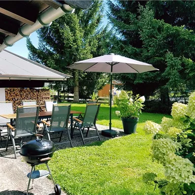 Ein schön gestalteter Garten mit einem Tisch und Stühlen unter einem Sonnenschirm. Im Vordergrund steht ein Grill und es umgeben weiße Hortensien.