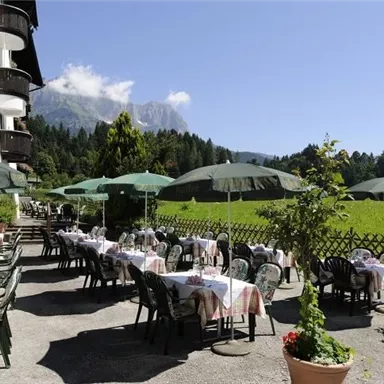 Ein einladendes Restaurant im Freien mit gedeckten Tischen und grünen Sonnenschirmen. Im Hintergrund sind blühende Wiesen und majestätische Berge zu sehen.