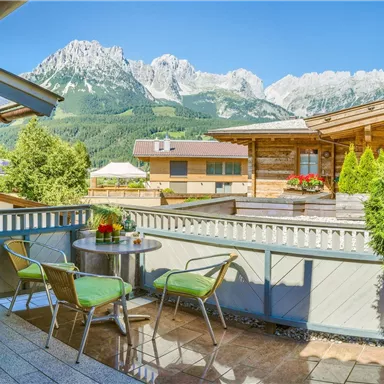 A sunny terrace with green chairs and a table, surrounded by mountains. The clear sky and nature create a relaxed atmosphere.