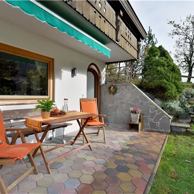 A cozy outdoor area with a table and two chairs. The space is surrounded by a green meadow and some trees.