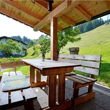 Eine gemütliche Holzveranda mit einem Tisch und Bänken. Im Hintergrund sieht man eine grüne Wiese und sanfte Hügel.
