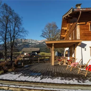 Ein schöner Holzbau mit einer Terrasse aus Holz und gemütlichen Stühlen. Im Hintergrund sind die Berge und Bäume zu sehen, die eine ruhige Landschaft schaffen.