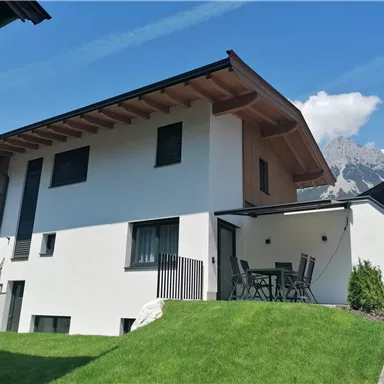 Ein modernes Haus mit zwei Etagen und einem großen Garten. Im Hintergrund sind Berge und ein blauer Himmel zu sehen.