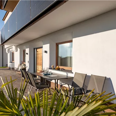 A modern terrace with seating and a dining table. In the background, you can see a light wall and a well-maintained garden.