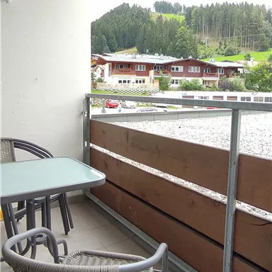 Ein Balkon mit einem Tisch und Stühlen. Im Hintergrund sind grüne Hügel und einige Holzgebäude sichtbar.
