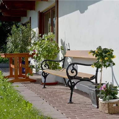 A sunny terrace with a wooden table and a metal bench. Surrounded by flowers and green grass, the space invites you to relax.