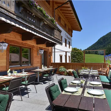 Eine sonnige Terrasse mit Blick auf die Berge. Es gibt Tische und Stühle, die zum Essen im Freien einladen.