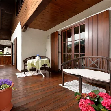 A cozy balcony with a wooden floor and two seating areas. Colorful flowers in pots give the space a cheerful atmosphere.