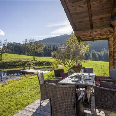 Eine gemütliche Terrasse mit einem Esstisch aus Rattan und Blick auf eine grüne Wiese und einen Teich. Im Hintergrund sind sanfte Hügel und Bäume zu sehen.