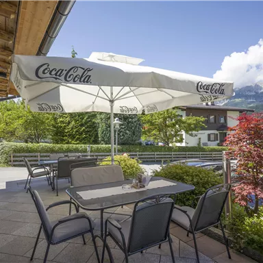 Ein gemütlicher Außenbereich mit Tischen und Stühlen unter einem großen Sonnenschirm. Im Hintergrund sind grüne Bäume und die Berge zu sehen.