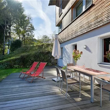 Eine gemütliche Terrasse mit Holzdielen und modernen Möbeln. Rote Liegestühle und ein Esstisch laden zum Entspannen im Freien ein.