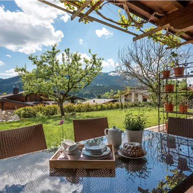 Ein einladender Essbereich im Freien mit einem Tisch und Stühlen. Im Hintergrund sind grüne Wiesen und Berge zu sehen.