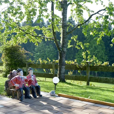 Drei Figuren sitzen auf einer Bank unter einem Baum. Im Hintergrund ist eine grüne Wiese mit einem Zaun und Bäumen sichtbar.