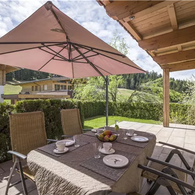 Eine gemütliche Terrasse mit einem Tisch, Stühlen und einem Sonnenschirm. Im Hintergrund sind grüne Wiesen und Holzgebäude zu sehen.