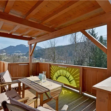 A cozy terrace with wooden furniture and a view of the mountains. The sun is shining, creating an inviting atmosphere.