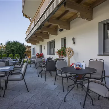 Eine gemütliche Terrasse mit Tischen und Stühlen, umgeben von Pflanzen. Die Sonne scheint und sorgt für eine angenehme Atmosphäre.