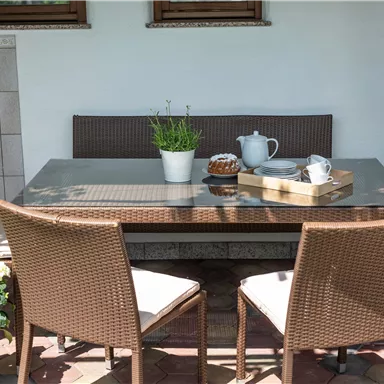 Ein gemütlicher Tisch mit Stühlen im Freien, umgeben von Pflanzen. Auf dem Tisch befinden sich Teekanne, Tassen und ein kleiner Kuchen.