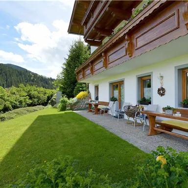 A cozy outdoor area with a large garden and seating. In the background, green hills and a blue sky can be seen.