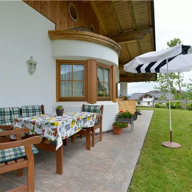 Eine gemütliche Terrasse mit einem dekorativen Tisch und Stühlen. Ein Sonnenschirm schützt vor der Sonne, umgeben von grünem Rasen und Pflanzen.