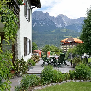 Ein schöner Garten mit einer Terrasse und Stühlen. Im Hintergrund sieht man beeindruckende Berge und eine Wolkenlandschaft.