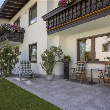 Ein modernes Haus mit einem gepflegten Garten. Es gibt gemütliche Stühle und Blumen an den Balkonen, umgeben von Bergen.
