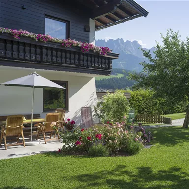 Ein hübsches Haus mit einem Balkon, auf dem Blumen blühen. Vor dem Haus befindet sich ein gemütlicher Garten mit einer Terrasse und Blick auf die Berge.