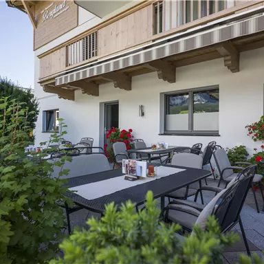 Eine einladende Terrasse mit Tischen und Stühlen, umgeben von Pflanzen. Im Hintergrund ist ein ruhiges Gebäude sichtbar.