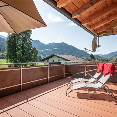 A sunny balcony with deck chairs and a sun umbrella. In the background, mountains and green forests can be seen.