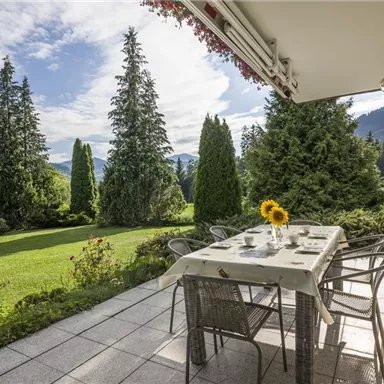 Eine einladende Terrasse mit einem Tisch und Stühlen. Im Hintergrund sind hohe Bäume und eine schöne Landschaft zu sehen.