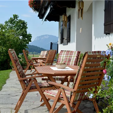 An inviting terrace with wooden furniture and colorful flowers. In the background, green meadows and mountains can be seen.