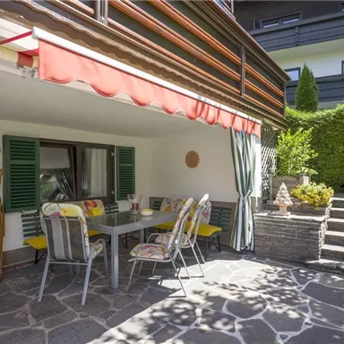 A cozy terrace with a dining table and chairs, surrounded by plants. The area has a sunny and inviting character.