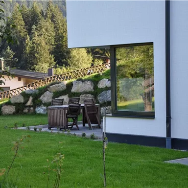 Ein modernes Haus mit großen Fenstern befindet sich in einer grünen Umgebung. Im Vordergrund sind Holzstühle und ein Tisch auf der Terrasse zu sehen.