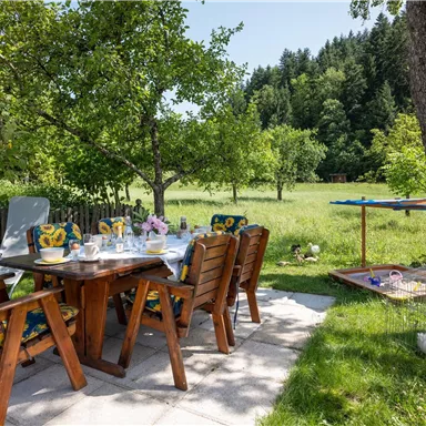 Ein einladender Essbereich im Freien mit Holzmöbeln und blühenden Pflanzen. Im Hintergrund erstreckt sich eine grüne Wiese mit Bäumen.
