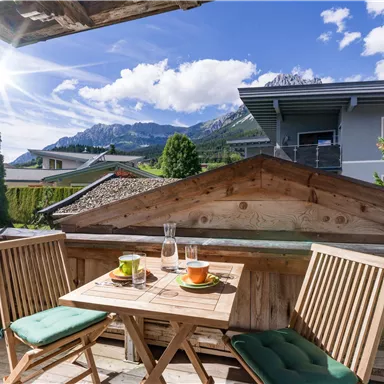 A cozy terrace with a view of the mountains. Two chairs and a table are decorated with drinks, while the sun shines.