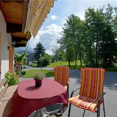 A cozy terrace with a round table and colorful chairs. In the background, trees and a quiet street can be seen.