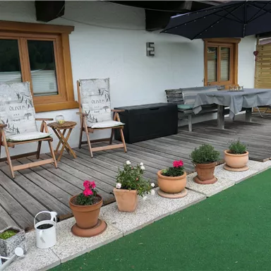 Eine gemütliche Terrasse mit zwei Liegestühlen, einem Tisch und einem Sonnenschirm. Blumenkübel und ein grüner Rasen schaffen eine einladende Atmosphäre.