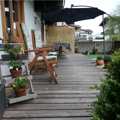 Eine gemütliche Terrasse mit Holzbohlen, Stühlen und blühenden Pflanzen. Ein Sonnenschirm bietet Schatten und es gibt einen Blick auf die Umgebung.