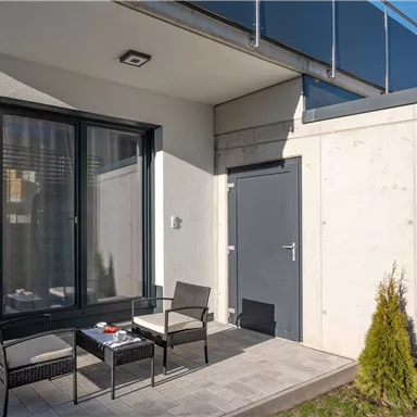 A modern terrace with two chairs and a table. In the background, there is a gray wall and large windows.