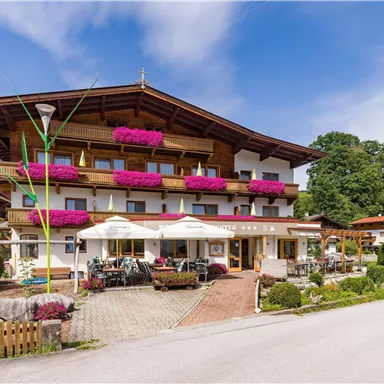 Ein charmantes Hotel mit blühenden Balkonpflanzen und einem einladenden Außenbereich. Umgeben von grüner Landschaft und Bergen, strahlt es eine gemütliche Atmosphäre aus.