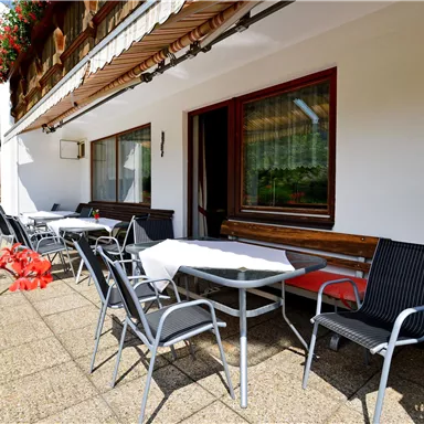 A terrace with tables and chairs, surrounded by flowers. The sun is shining and it is an inviting outdoor area.
