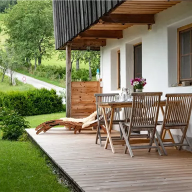 A cozy outdoor area with a wooden table and chairs on a porch. In the background, green meadows and trees are visible.
