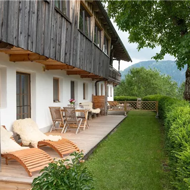A cozy outdoor space with wooden lounge chairs and a table. In the background, green hills and a beautiful tree can be seen.