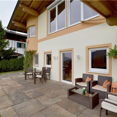 Eine gemütliche Terrasse mit modernen Möbeln und einem gepflegten Garten. Im Hintergrund sind Häuser und Grünpflanzen sichtbar.