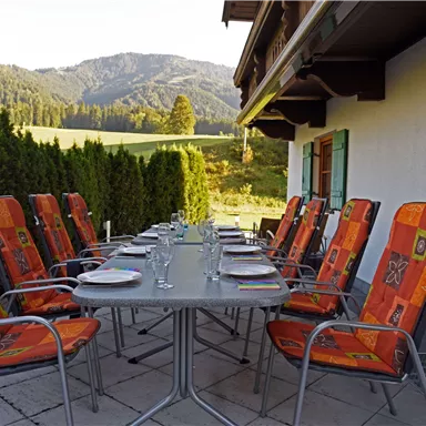 A well-kept dining table on a terrace with orange chairs, surrounded by green trees and hills. In the background, mountains can be seen, forming a beautiful landscape.