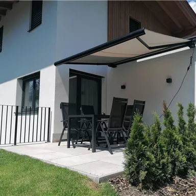 Eine moderne Terrasse mit einem Tisch und Stühlen unter einem Sonnenschirm. Der Bereich ist von einem Rasen und kleinen Sträuchern umgeben.