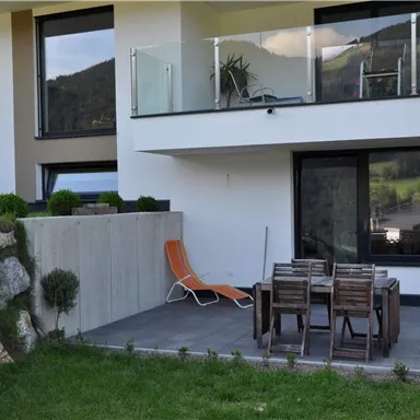 Eine moderne Terrasse mit einem Tisch und Stühlen aus Holz. Im Hintergrund ist ein elegantes Haus mit großen Fenstern und einem Balkon zu sehen.