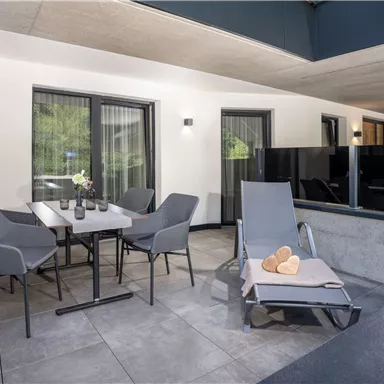 A modern, inviting patio with a table, chairs, and a lounge chair. The surroundings are adorned with flowers, creating a cozy atmosphere.