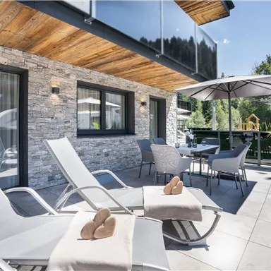 A modern terrace with loungers and a sun umbrella. In the background, you can see a well-kept garden and a playground.