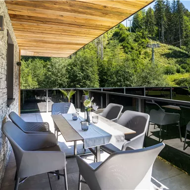 A modern balcony with elegant furniture and a table. The surroundings feature lush greenery and a clear sky.